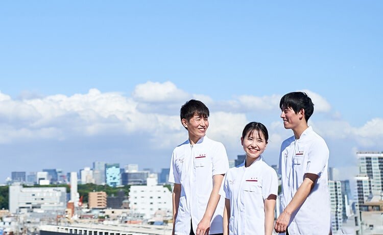 アイン薬局のユニフォームを着たフレッシュな薬剤師4名が笑顔で会話しながら歩いている。背景には青い空と白い雲、東京の街が写っている。