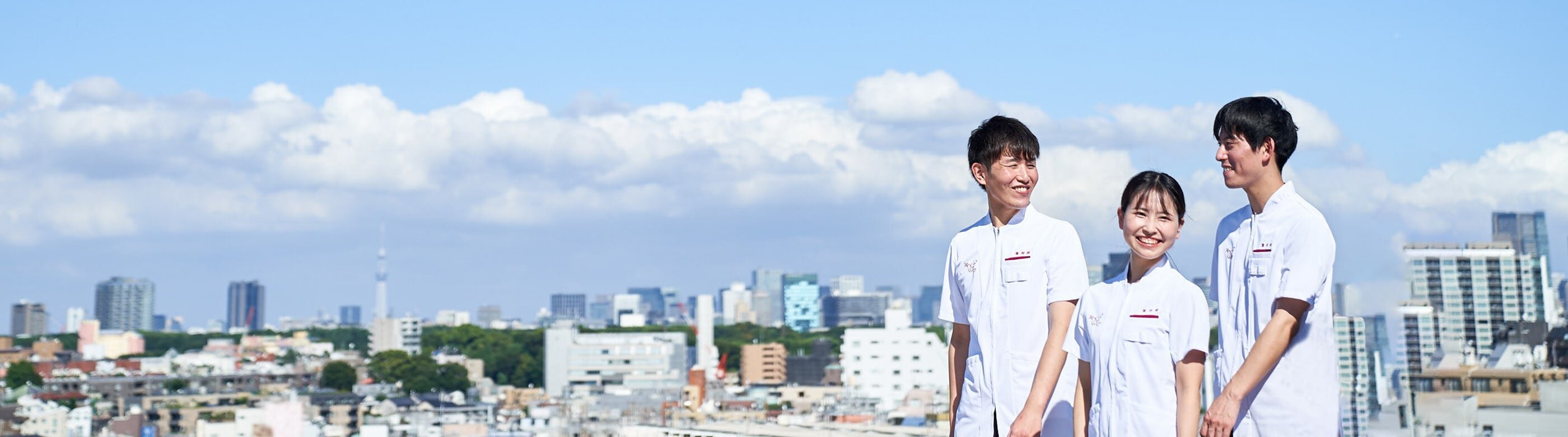 アイン薬局のユニフォームを着たフレッシュな薬剤師4名が笑顔で会話しながら歩いている。背景には青い空と白い雲、東京の街が写っている。