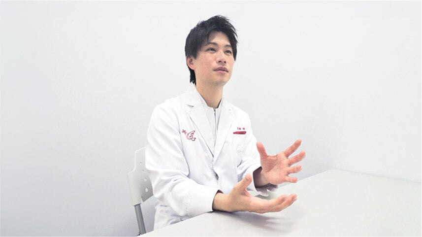 白衣を着用した人物。左胸にアインの赤いマーク。何かを説明している手ぶり。