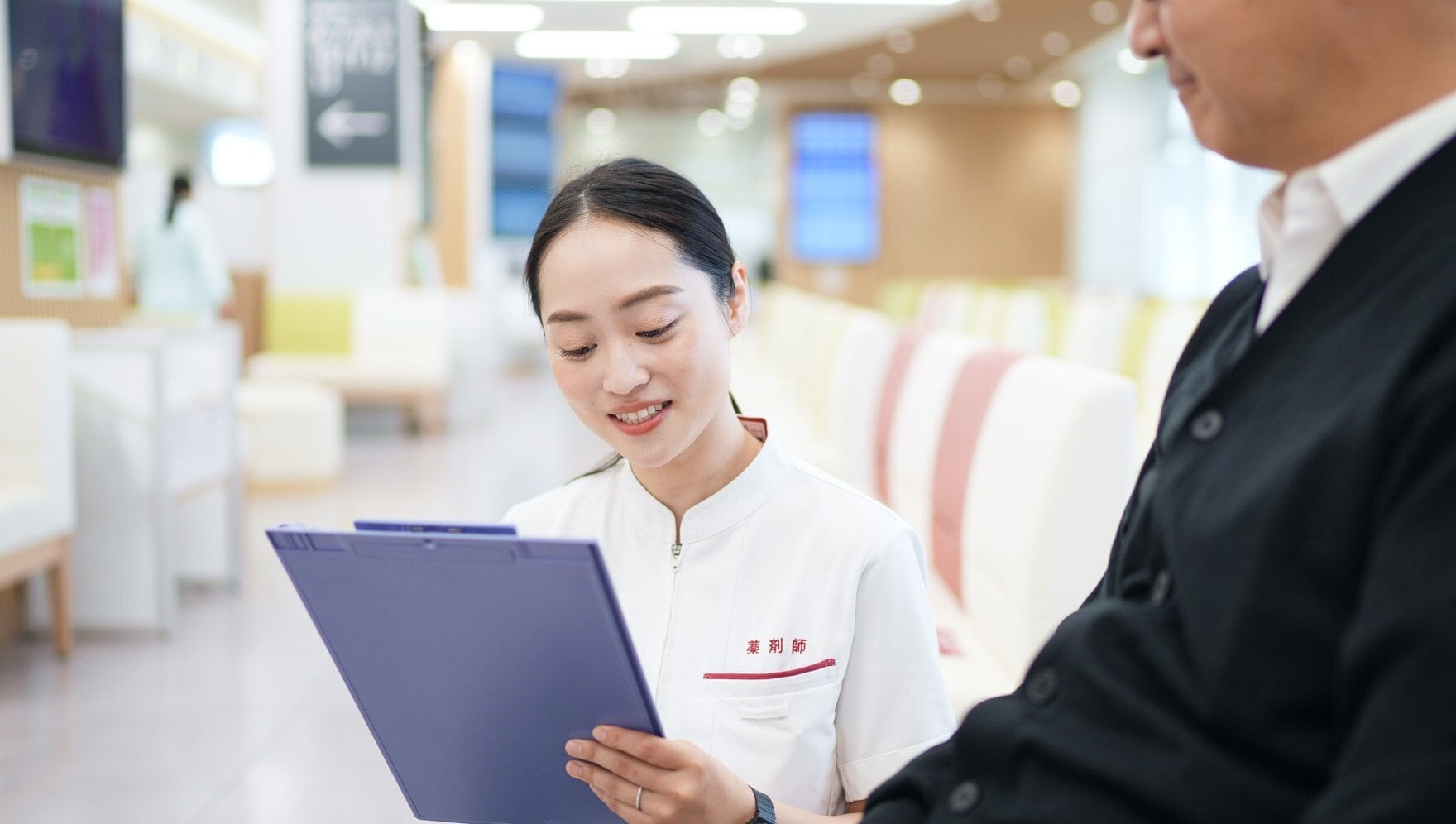 「薬剤師」と書かれた白いユニフォームの女性がバインダーを見て、隣に立つ人物。