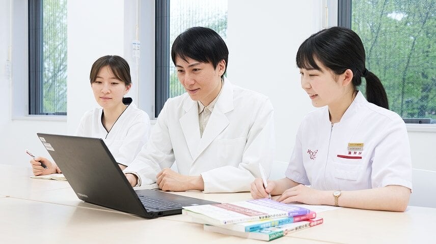 白衣の男性と2人の女性が机で作業している。男性はノートパソコンを見て、女性たちは本や書類に書き込んでいる。1人の名札には「薬剤師」。