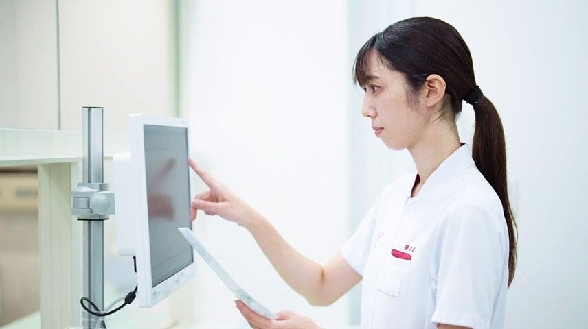 白いユニフォームのポニーテールの女性が、モニターに指で触れ、左手に書類を持つ。