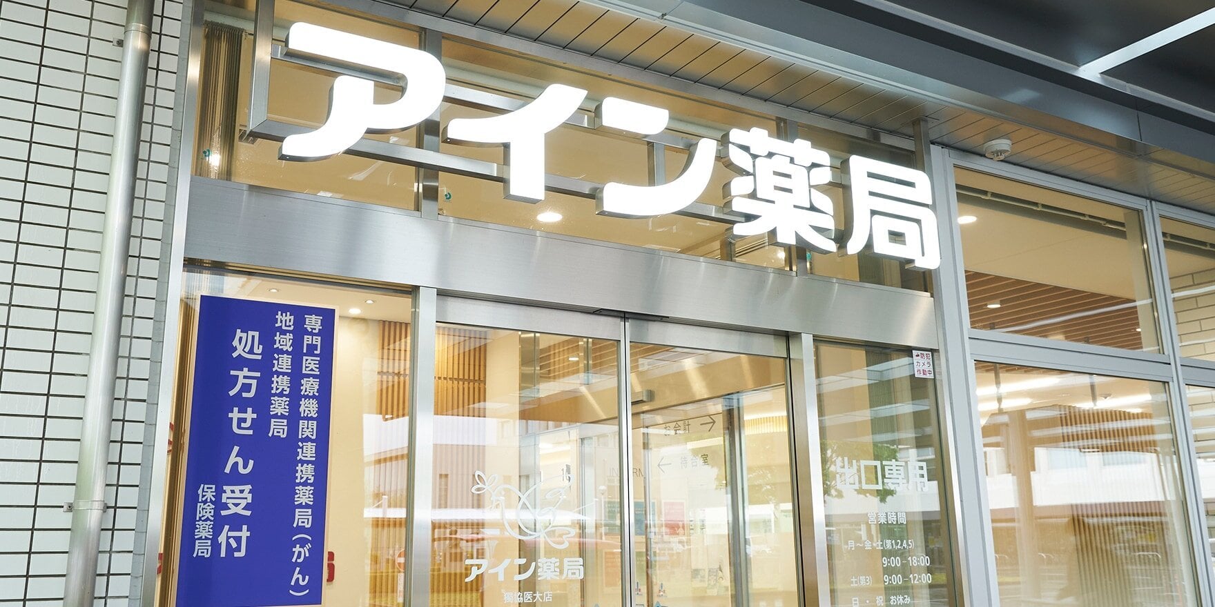 アイン薬局の入り口。専門医療機関連携薬局（がん）、地域連携薬局、処方せん受付の表示と営業時間案内がある。